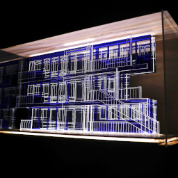 3D geometric acrylic model, rectangular windows and blue features, in a glass case with wooden back under LED lights.