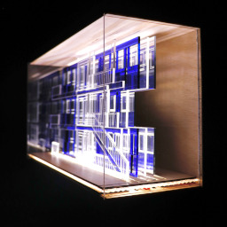 3D geometric acrylic model, rectangular windows and blue features, in a glass case with wooden back under LED lights.