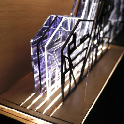 3D geometric acrylic model, angular features and blue tones, in a glass case with wooden back under LED lights.