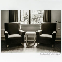 two armchairs sitting on wooden floorboards, small side-table in the centre, in front of a window, greenery outside, black and white photograph.