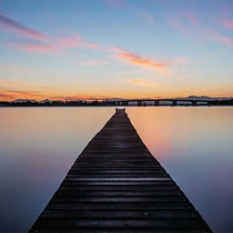 long boardwalk heading out into the water, sunset sky with blues, yellows and pinks.