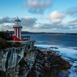 red and white lighthouse sitting on rocky cliffe, below crashing waves on rocks and a cloudy sky above.