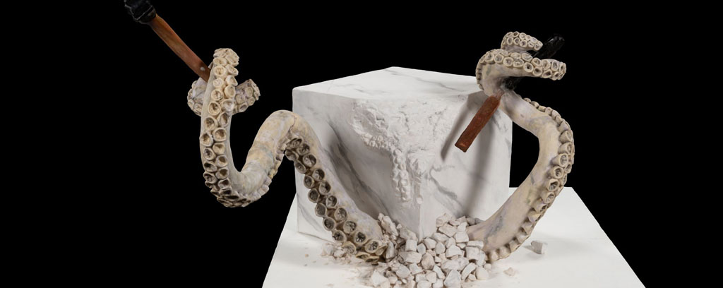 marble block sculpture with two tenticles breaking out of the rock holding hammers on a white base.