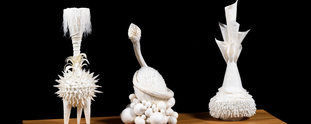 three tall white ceramic sculptures with curved lines and textural detail sitting on wooden shelf.