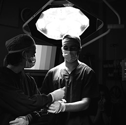 black and white photo of a surgeon standing under a spot light being handed an instrument