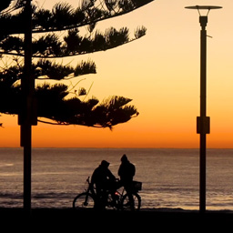 Still shot of silhouettes of two people on bikes by the ocean with an orange sunrise in the background.